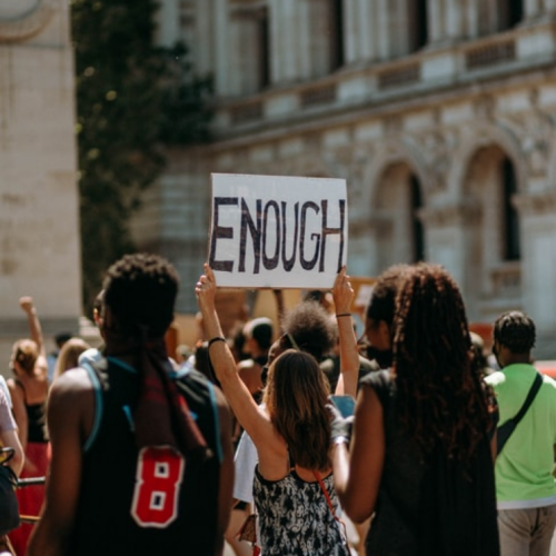 A woman at a protest holding a sign reading 'enough.' She stands in a crowd of other protesters.